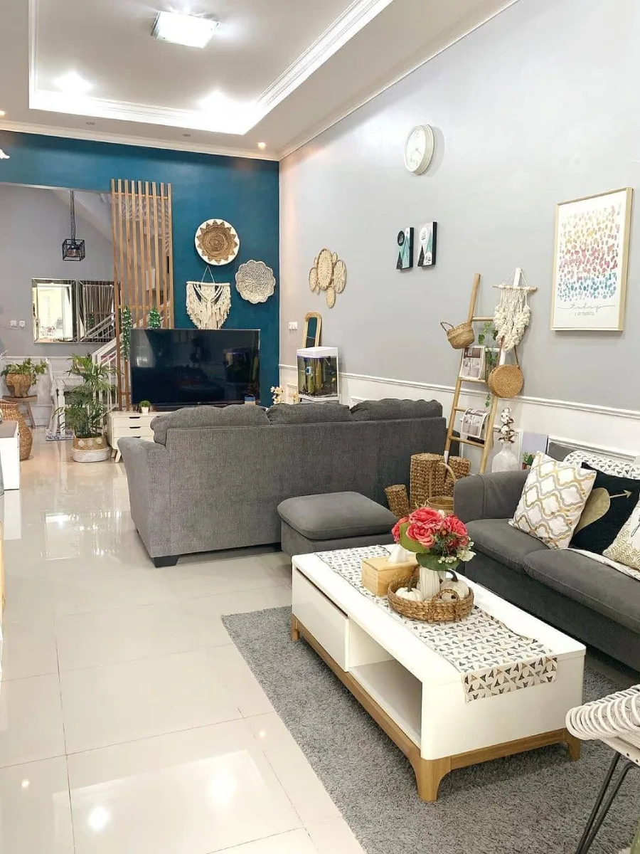 Ruang tamu minimalis dengan dinding aksen teal, dekorasi dinding rotan, sofa abu-abu gelap, dan meja kopi putih yang rapi.