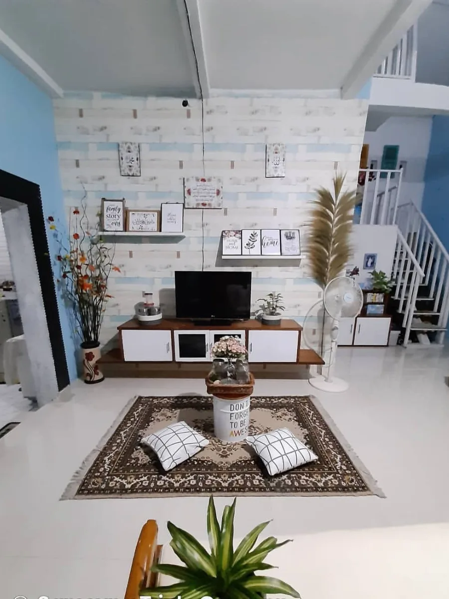 Desain ruang keluarga lesehan dengan karpet permadani, bantal lantai, backdrop TV motif bata putih, dan dekorasi bunga kering yang estetik.