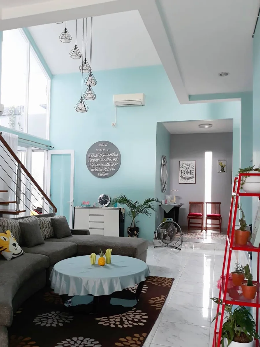 Ruang keluarga plafon tinggi (high ceiling) dengan cat dinding warna mint, lampu gantung geometris, dan sofa sudut L warna abu-abu.