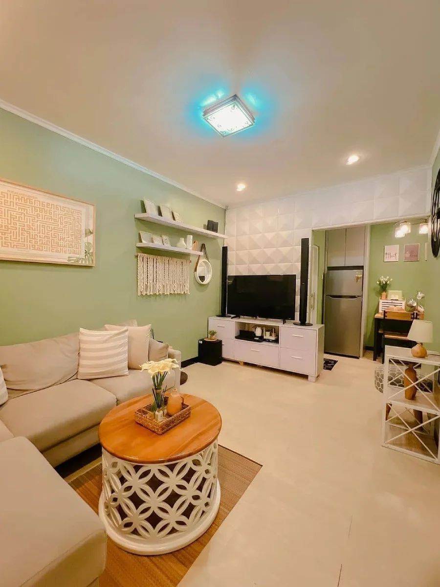 Ruang keluarga estetik dengan dinding hijau sage, meja tamu kayu bulat motif laser cut, dan panel dinding 3D putih di belakang TV.