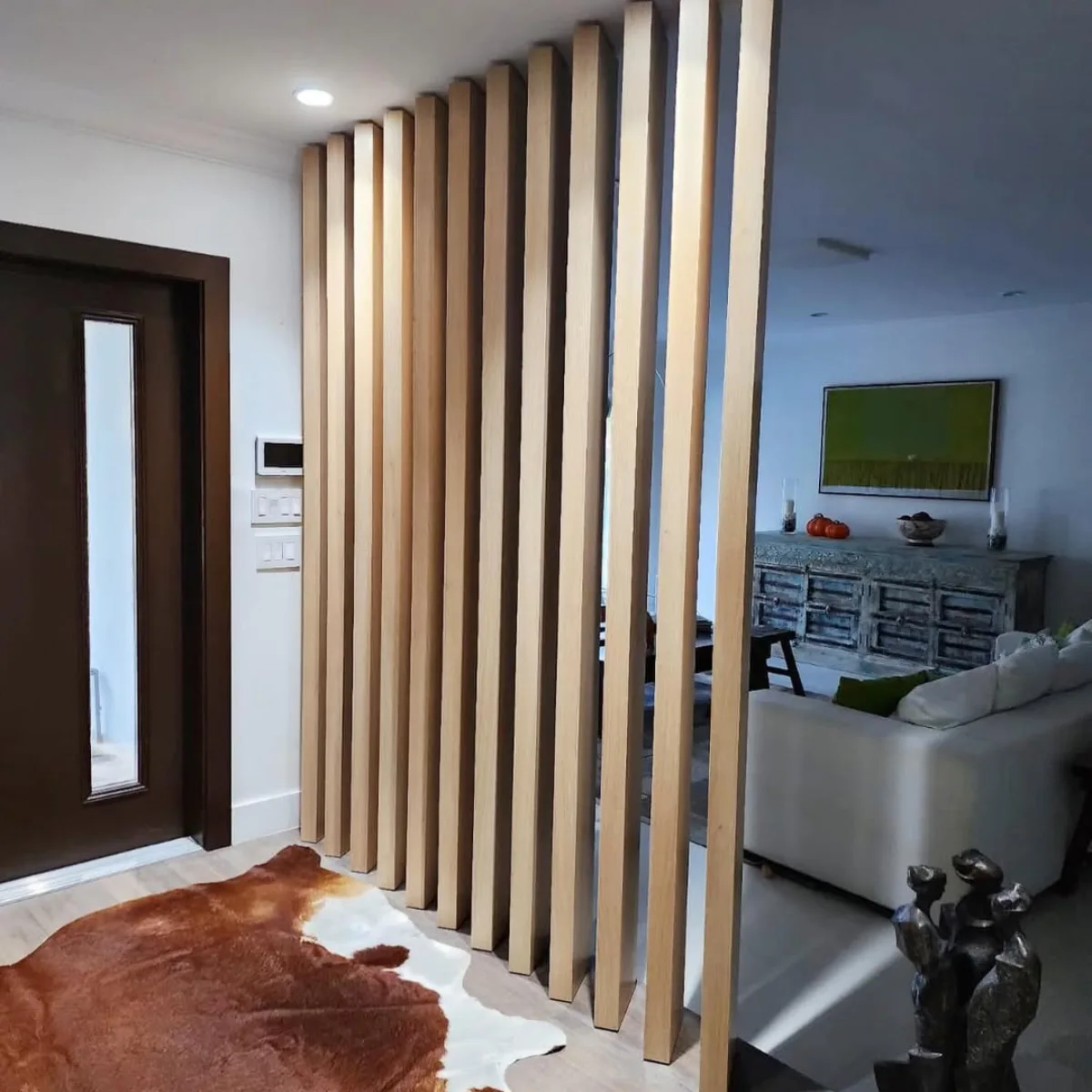Sekat ruangan yang terbuat dari jajaran bilah kayu vertikal tebal berwarna cokelat muda alami, memisahkan area pintu masuk berkarpet kulit sapi dengan ruang keluarga di dalamnya.