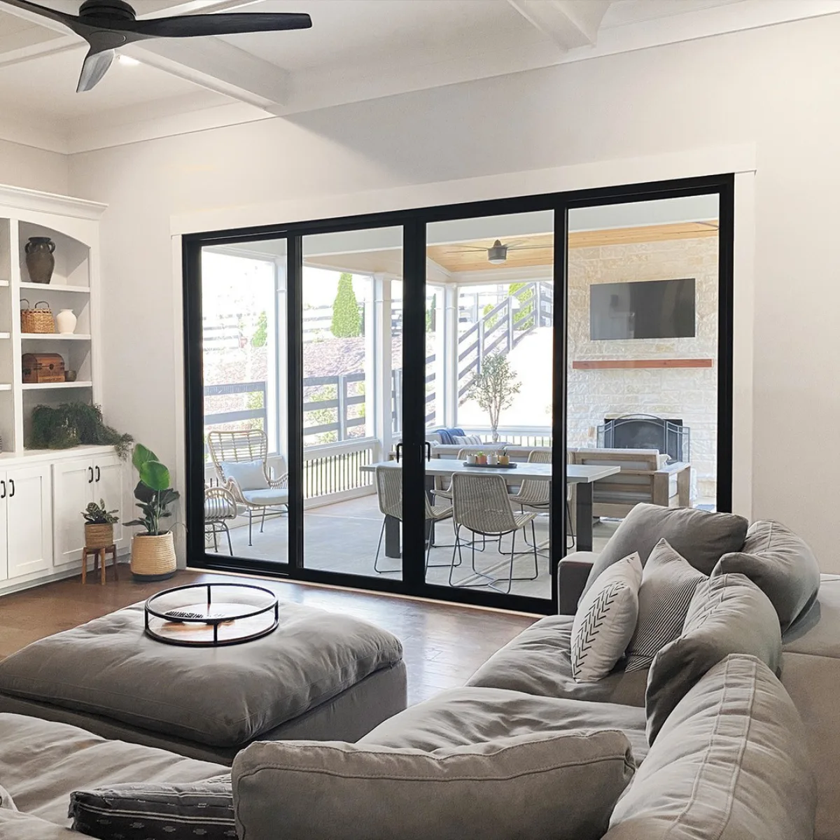 Pintu geser kaca besar berbingkai hitam minimalis yang memisahkan area ruang tamu dengan sofa abu-abu dan area teras luar ruangan tempat makan.
