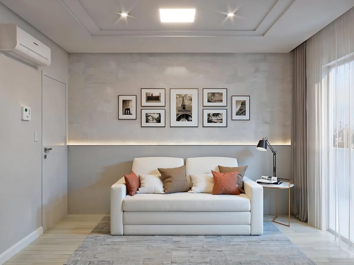 Area santai di home office dengan sofa putih, bantal warna bumi, lampu sudut, dan dinding bertekstur semen dengan susunan bingkai foto (gallery wall).