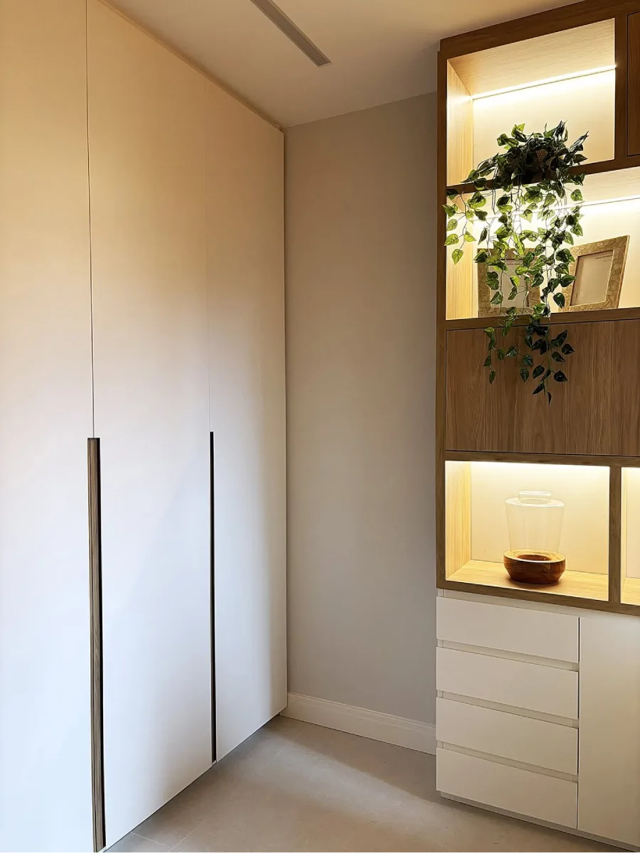 Detail kabinet lemari putih minimalis yang menyatu dengan rak kayu vertikal, dihiasi tanaman rambat artifisial dan pencahayaan hangat.