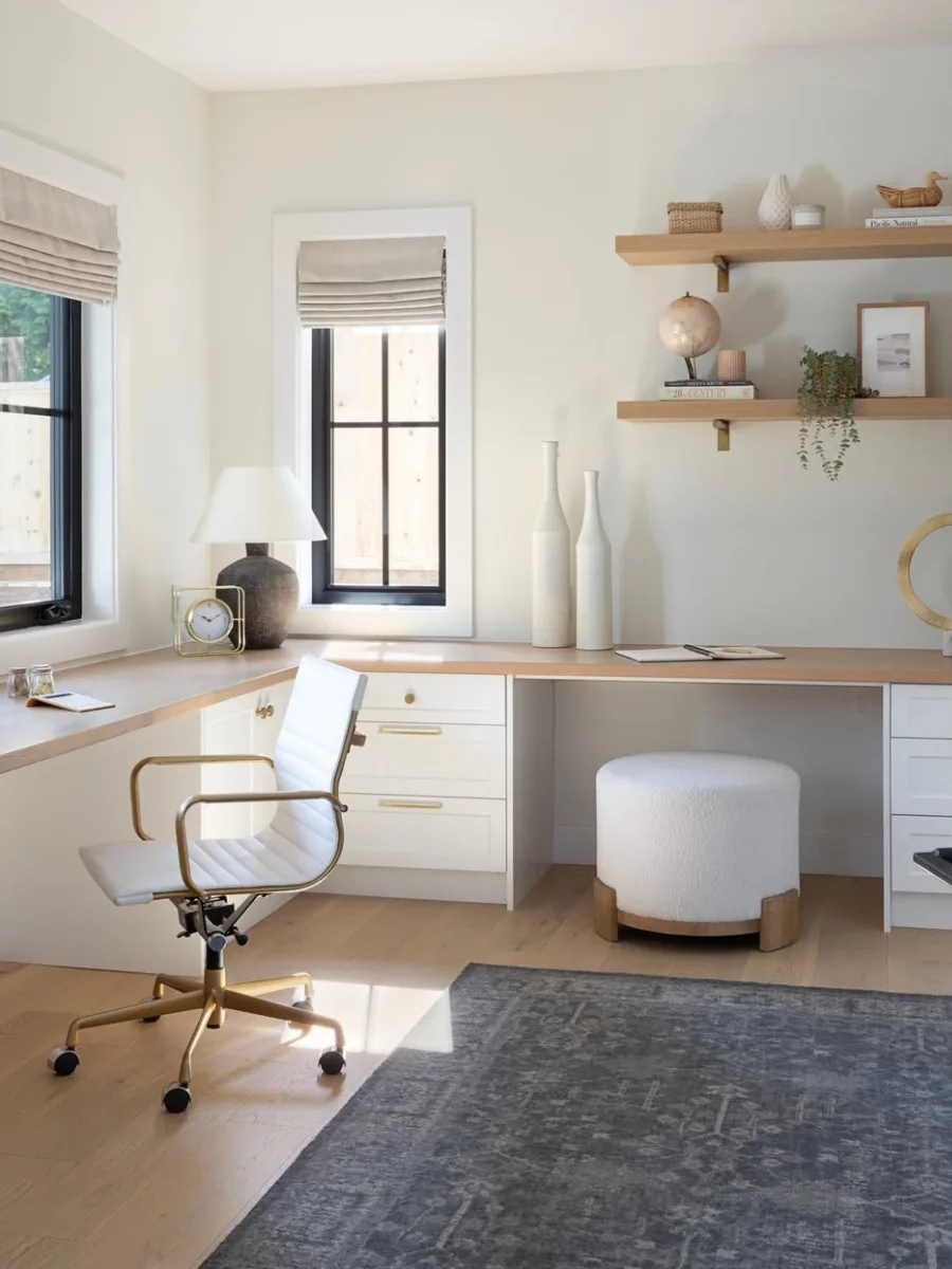 Meja kerja sudut bentuk L (L-shape) warna putih di dekat jendela dengan cahaya alami maksimal, dilengkapi kursi putih beraksen emas dan rak dinding kayu.