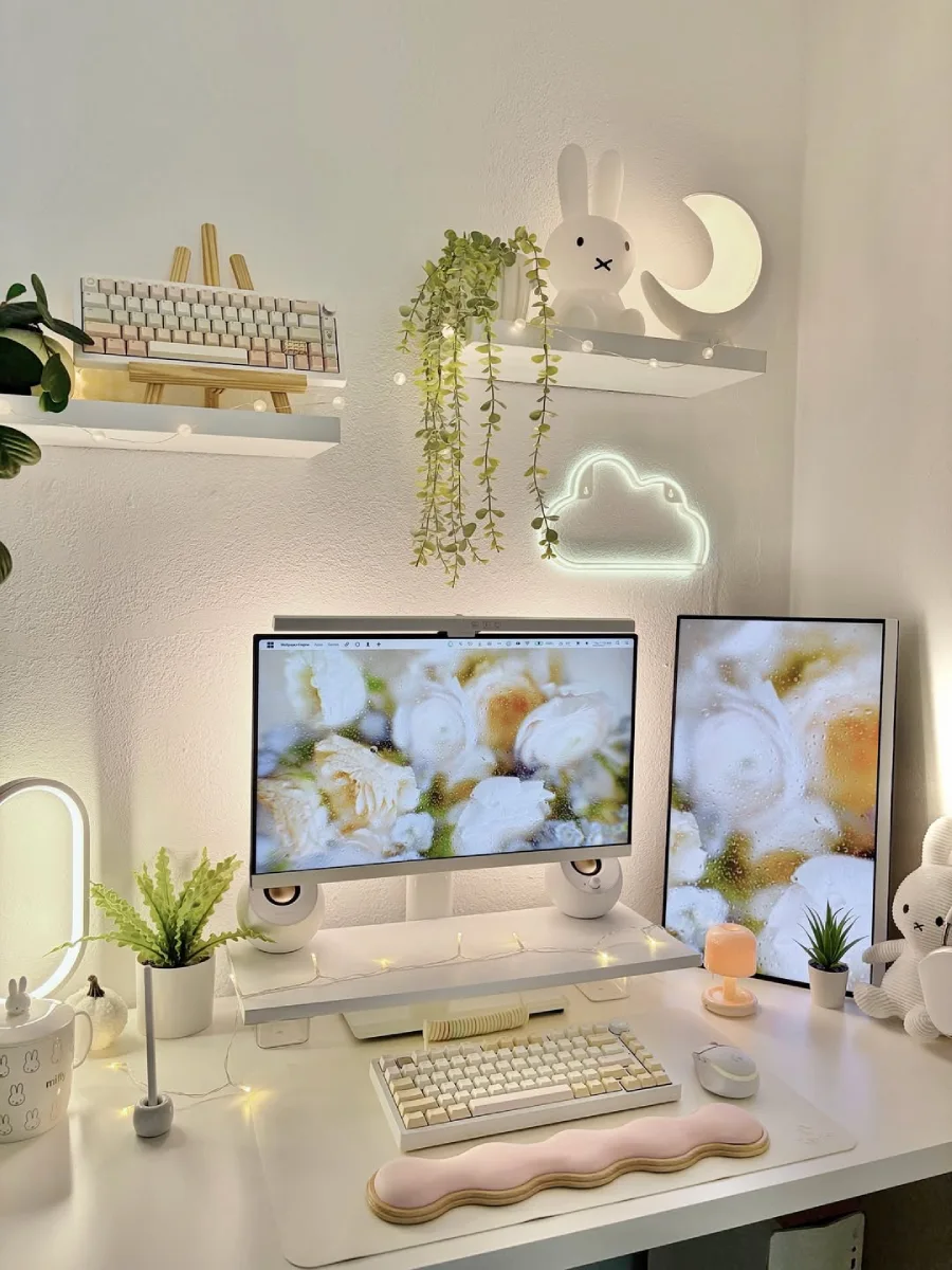 Sudut meja kerja bernuansa putih bersih memperlihatkan keyboard mekanik dengan aksen pink yang dipajang layaknya lukisan di atas stand kayu mini, ditemani tanaman hias kecil yang menyegarkan.