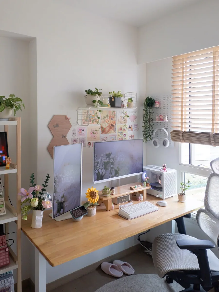 Sudut pandang lebih luas dari meja kerja bernuansa kayu dan putih, memperlihatkan rak putih berisi pajangan bunga di sisi kiri, jendela dengan tirai bambu di sisi kanan, serta kursi ergonomis abu-abu.