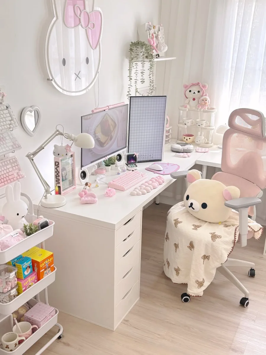 Meja kerja L-shape bertema pink pastel dan putih dengan lampu neon Miffy di dinding, dua monitor, keyboard pink dengan wrist rest bentuk awan, dan trolley penyimpanan susun di sebelah kiri.