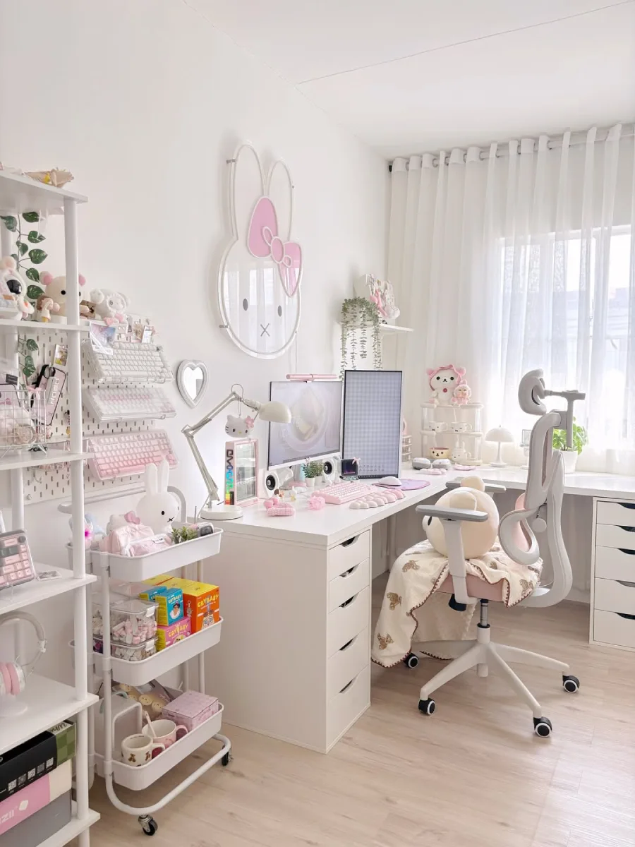 Tampak samping meja kerja pink pastel, memperlihatkan rak dinding pegboard putih yang menggantung keyboard mekanikal, kursi kerja pink dengan bantal Rilakkuma, dan jendela dengan gorden putih tipis.