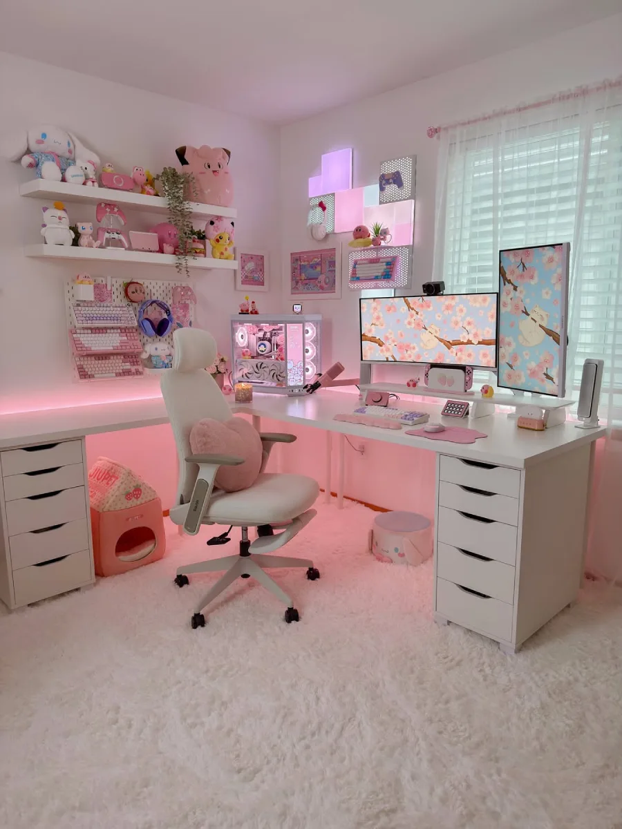 Setup meja gaming wanita bernuansa serba pink neon dengan karpet bulu putih tebal, dua monitor ber-wallpaper bunga sakura, casing PC berlampu RGB, dan rak dinding penuh boneka Kirby serta Pokemon.
