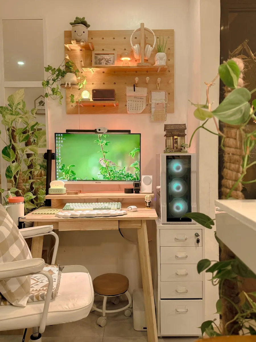 Meja kerja cozy bernuansa alam dengan dinding pegboard kayu, layar monitor ber-wallpaper burung hijau, tanaman hias rambat, dan casing PC dengan kipas menyala diletakkan di atas laci putih terpisah.