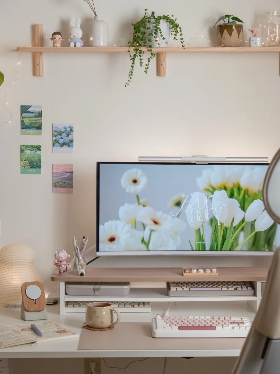 Detail area tengah meja kerja minimalis krem, memperlihatkan layar monitor ber-wallpaper bunga putih, kartu pos estetik yang ditempel di dinding, keyboard mekanikal, dan gelas keramik motif bunga.