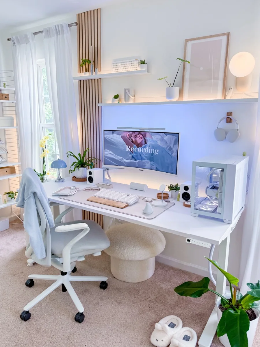 Meja kerja estetik berwarna putih di dekat jendela dengan panel dinding kayu terang, rak melayang berisi tanaman hias kecil, monitor lengkung besar, kursi ergonomis putih dengan sweter biru tersampir, dan penyangga kaki berbentuk jamur yang menggemaskan.