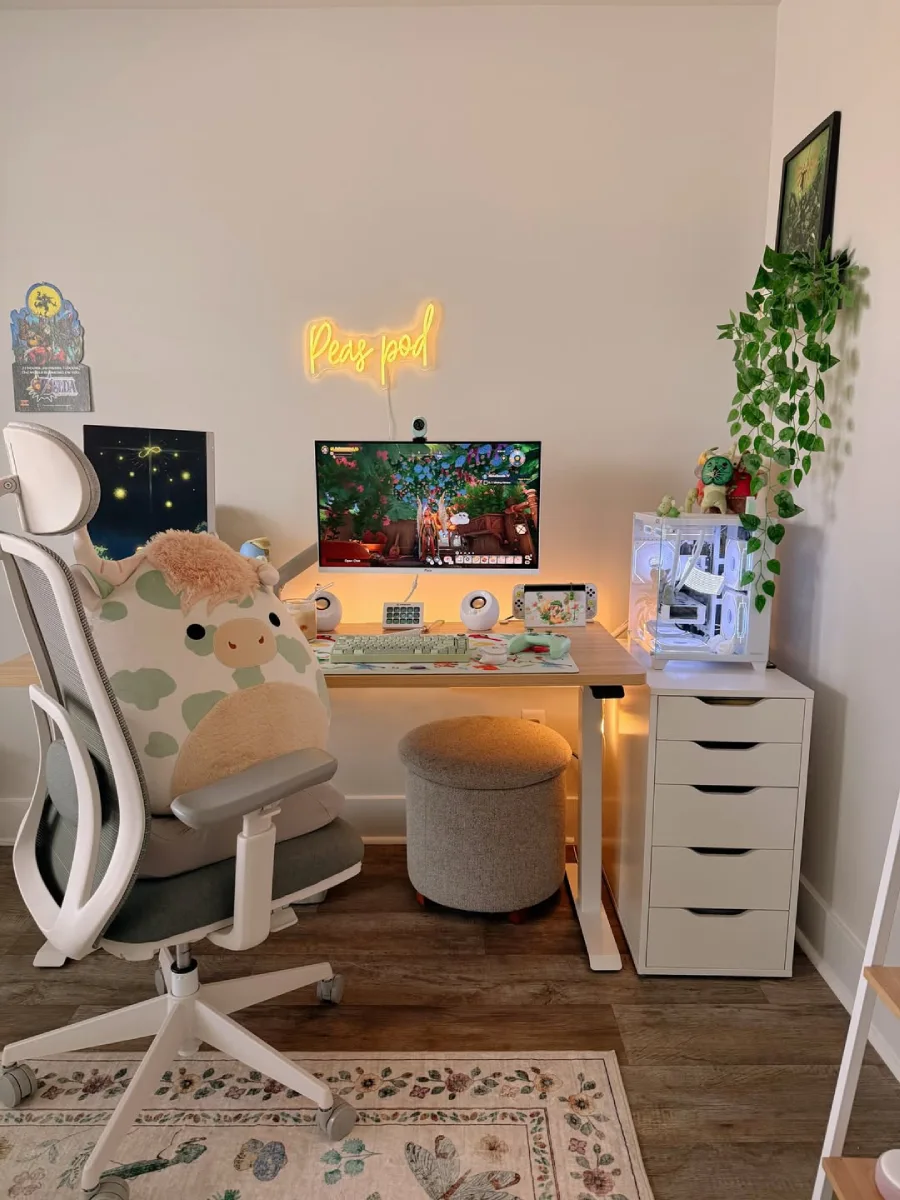 Setup meja kerja berbahan kayu yang hangat dan cozy, dilengkapi laci putih berlapis, lampu neon kuning bertuliskan 'Peas pod', tanaman gantung, dan kursi ergonomis abu-abu yang dihiasi boneka plushie sapi berukuran besar.