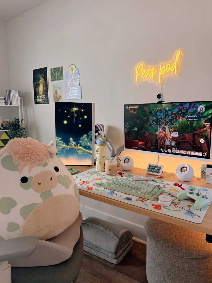 Tampilan jarak dekat meja kerja kayu bergaya gamer, memperlihatkan layar monitor utama yang menampilkan sebuah game, layar vertikal dengan wallpaper pemandangan malam, desk mat bermotif lucu, keyboard hijau pastel, dan segelas es kopi.