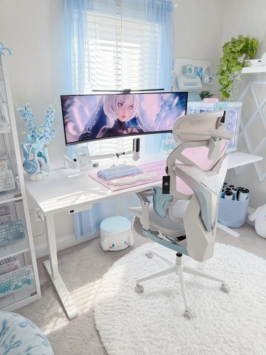 Meja kerja bernuansa kawaii dengan dominasi warna biru pastel dan pink. Menampilkan monitor ultrawide dengan wallpaper karakter anime, kursi gaming warna pink dan putih, keyboard biru, serta karpet bulu putih bundar di lantai.