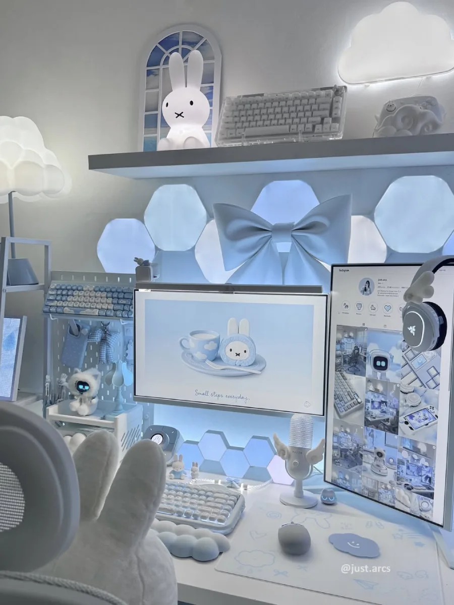 Detail dari meja kerja bertema biru awan, menampilkan layar monitor dengan gambar kelinci 3D di dalam cangkir teh, microphone putih bersayap kecil, desk mat motif awan, dan layar vertikal yang membuka profil Instagram.