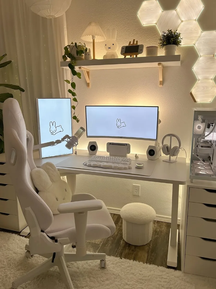 Meja kerja minimalis serba putih dengan nuansa cahaya kuning yang hangat (warm light). Dilengkapi kursi gaming putih berbantal kelinci, PC serba putih, laci di kanan-kiri meja, panel lampu heksagonal motif kayu, dan layar monitor bergambar Miffy.