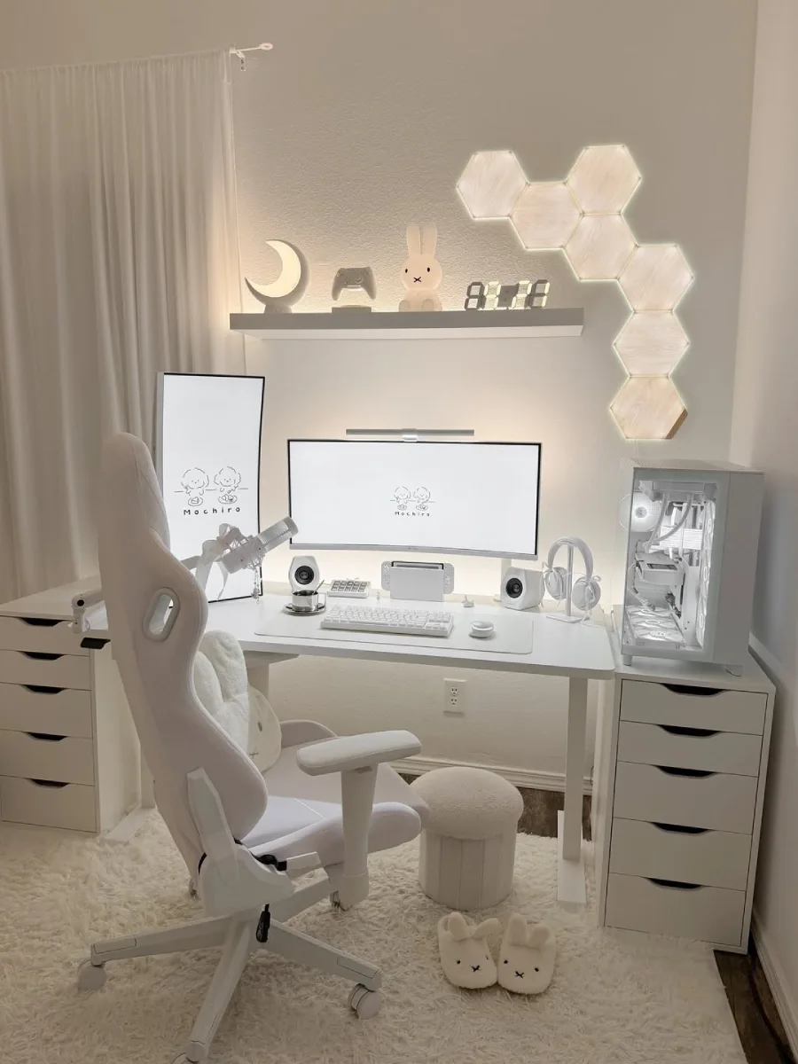 Setup serba putih yang rapi dengan layar monitor menampilkan wallpaper karakter kecil bertuliskan 'Mochiro'. Di atasnya terdapat rak melayang yang dihiasi lampu bulan, lampu Miffy, controller game, dan jam digital menyala terang.
