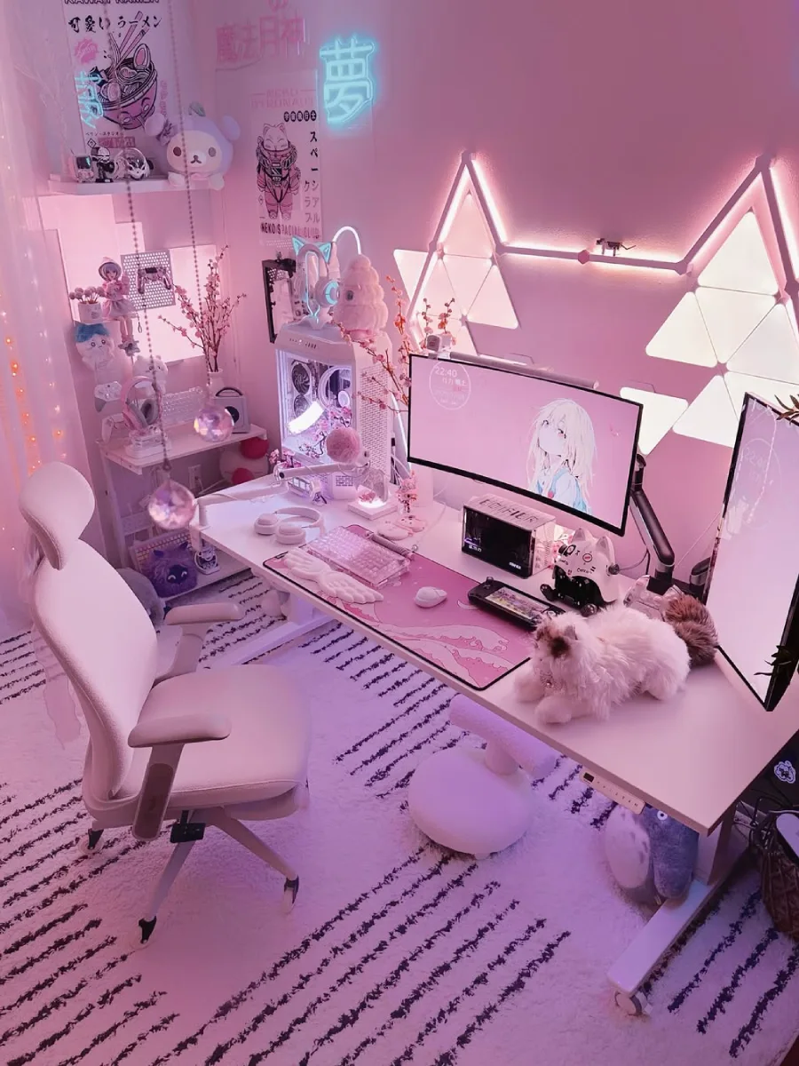 Setup meja kerja bernuansa pink pastel dan putih yang feminin. Dinding dihiasi lampu LED yang dibentuk menyerupai telinga kucing besar. Di meja terdapat desk mat pink motif ombak, monitor, PC case putih dengan lampu pink, dan boneka kucing putih berbulu lebat.