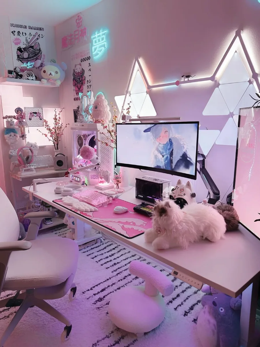 Tampilan sedikit lebih dekat dari meja kerja anime bernuansa pink. Memfokuskan pada layar monitor dengan wallpaper karakter anime perempuan, keyboard mekanikal bernuansa pink-putih, mouse, dan boneka kucing lucu yang berbaring santai di atas meja.