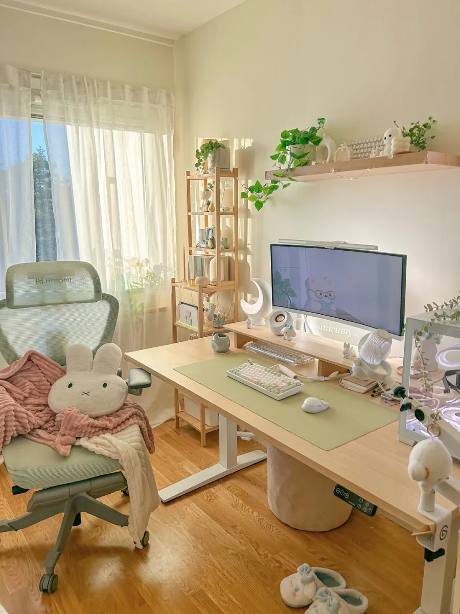 Meja kerja minimalis dan natural dengan perpaduan elemen kayu terang dan warna sage green. Tersorot cahaya matahari alami dari jendela, memperlihatkan kursi ergonomis hijau mint, meja kayu, tanaman hias, dan boneka kepala Miffy di atas kursi.