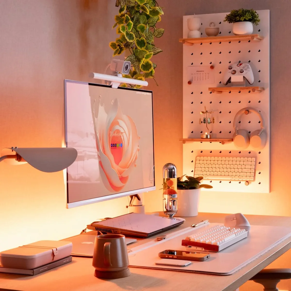 Sudut samping meja kerja estetik yang rapi, memperlihatkan detail pegboard putih di dinding yang digunakan untuk menggantung controller console, headphone, rak kecil, dan keyboard mekanik.