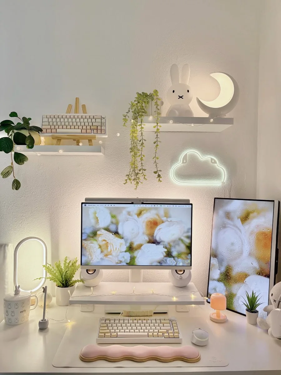 Meja kerja serba putih yang dreamy dengan monitor ganda menampilkan wallpaper bunga, lampu karakter Miffy, lampu bentuk bulan, neon awan, fairy lights, dan wrist rest bergelombang warna pink.
