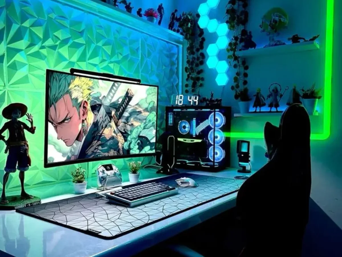 Setup meja gamer modern dengan lampu LED hijau biru, monitor menampilkan karakter anime, PC RGB transparan, panel dinding 3D, dan rak koleksi figure di sudut ruangan.
