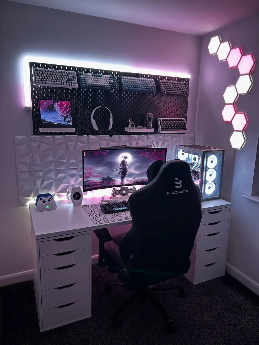 Setup gaming bernuansa pink dan ungu dengan meja putih, kursi gaming hitam bermerek Blacklyte, monitor melengkung ber-wallpaper samurai, PC putih bercahaya RGB, pegboard hitam yang memajang koleksi keyboard mekanik, dan lampu dinding heksagonal.