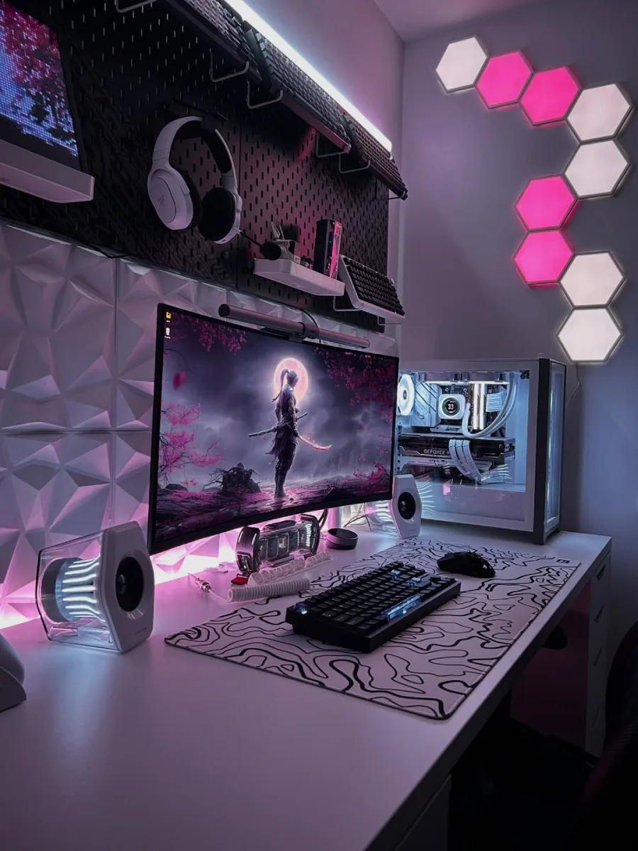 Tampilan jarak dekat setup gaming di meja putih dengan mousepad bermotif topografi, monitor melengkung menampilkan siluet samurai di bawah bulan, speaker putih, PC transparan bercahaya putih, dan pegboard hitam di dinding.