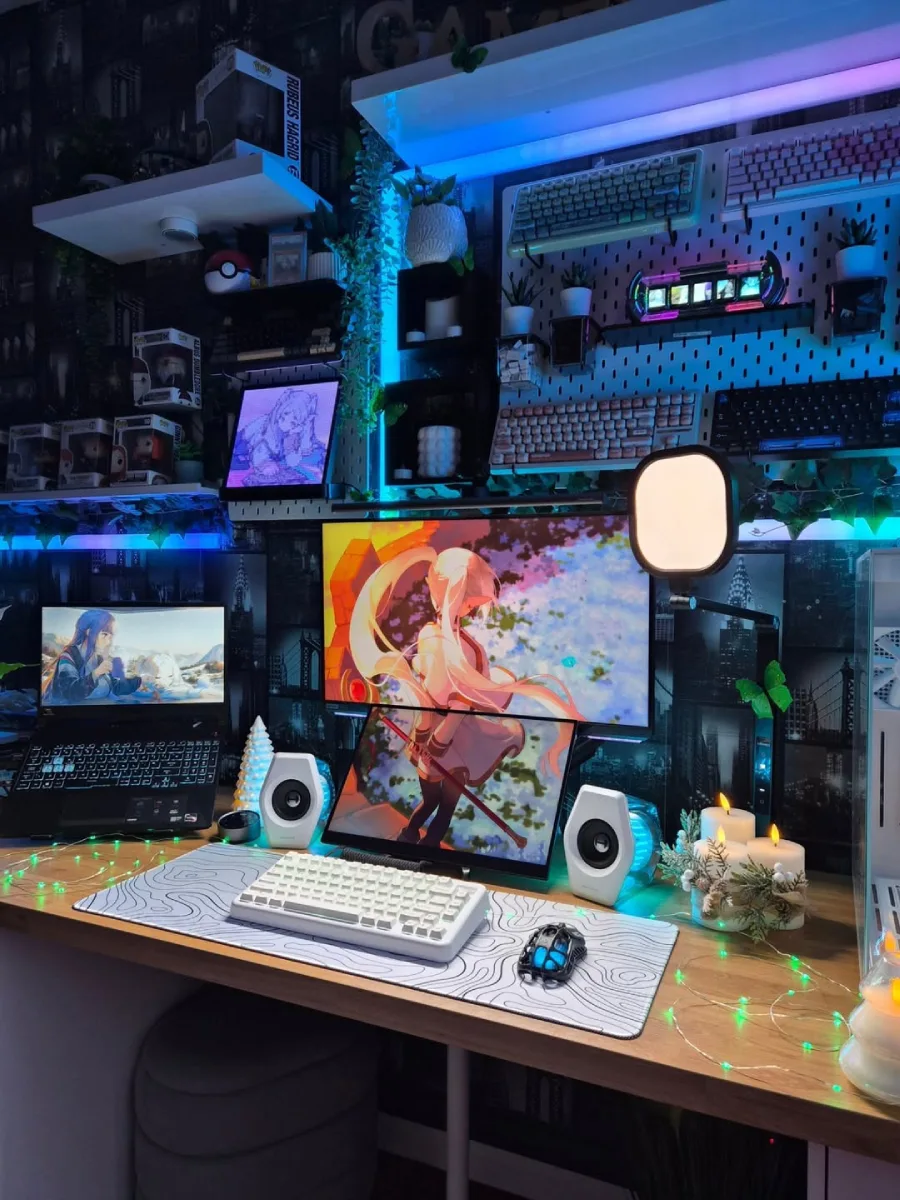 Sudut lain dari setup meja cozy dengan pencahayaan biru neon, menampilkan monitor bertumpuk ber-wallpaper karakter anime memegang pedang, speaker putih, lilin LED hias, dan dinding yang dihiasi wallpaper siluet gedung perkotaan.