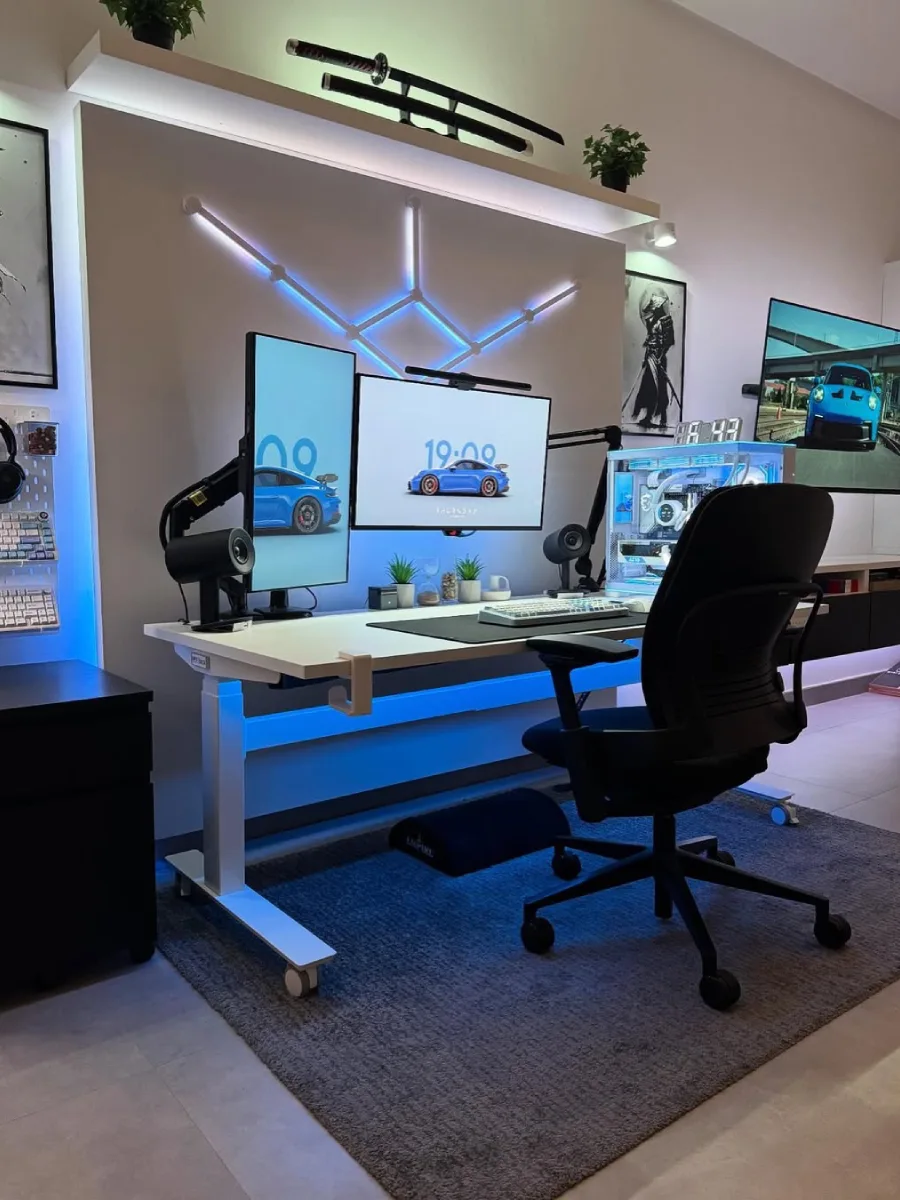 Sudut samping dari setup minimalis putih-biru, memperlihatkan kursi ergonomis hitam, meja standing desk putih, monitor dengan wallpaper mobil sport biru, lampu dinding geometris, dan dekorasi katana di bagian atas.