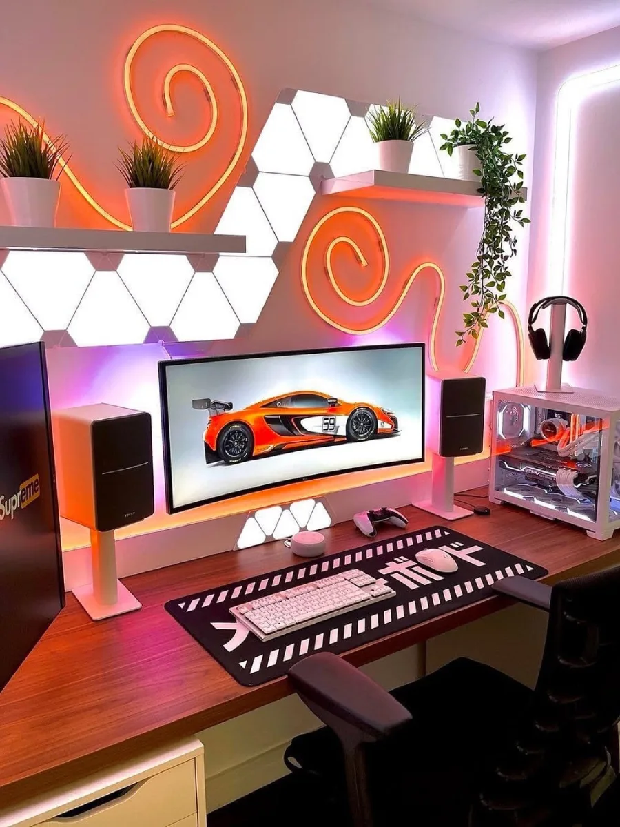 Setup gaming cerah dengan pencahayaan oranye dan pink. Terdapat monitor lebar ber-wallpaper mobil McLaren oranye, hiasan lampu neon fleksibel yang melingkar di dinding bersama panel heksagonal putih, rak dengan tanaman hias kecil, dan desk mat hitam bermotif huruf Jepang.
