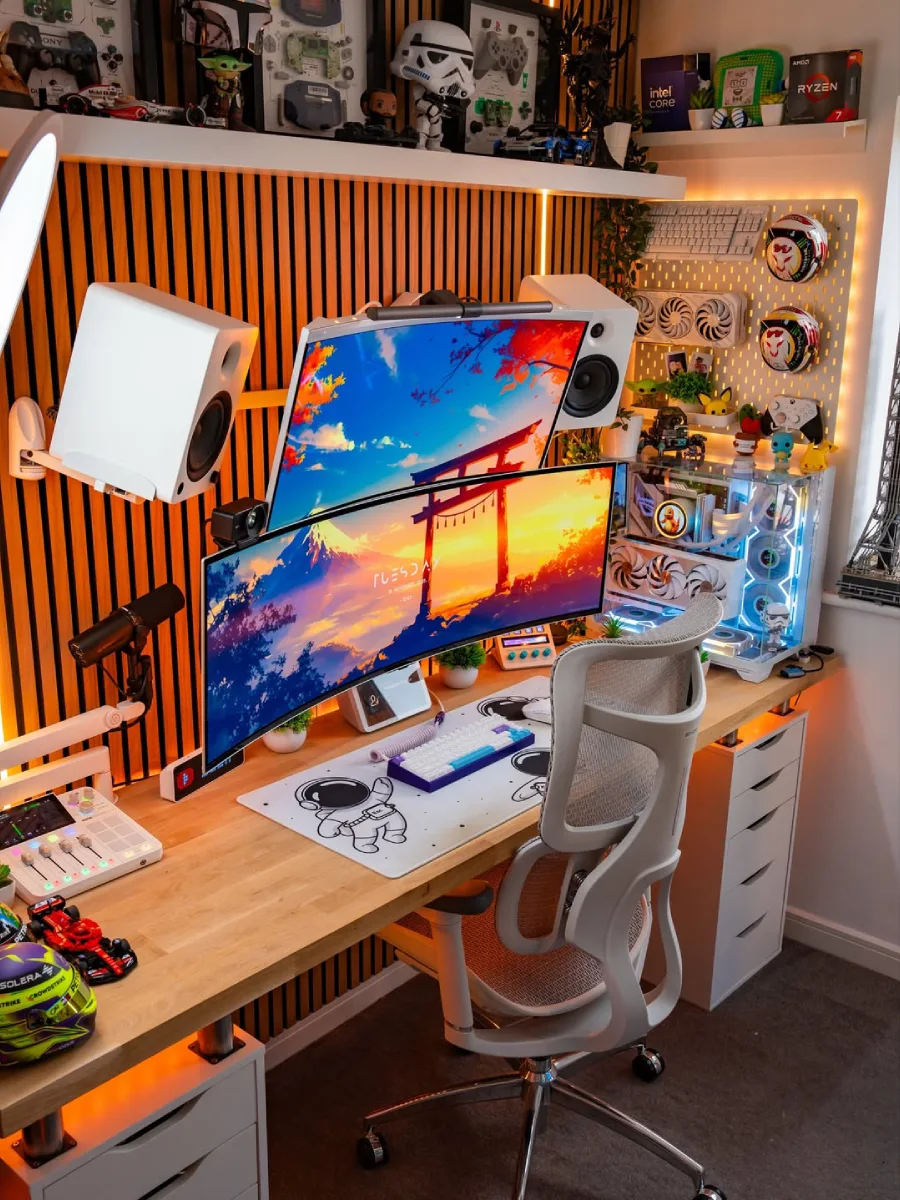 Tampilan dekat setup PC berdinding panel kayu. Fokus pada dua monitor melengkung yang ditumpuk menampilkan wallpaper pemandangan anime. Terdapat desk mat putih bergambar astronot, keyboard mekanikal, speaker putih, dan pegboard berisi helm miniatur di dinding kanan.