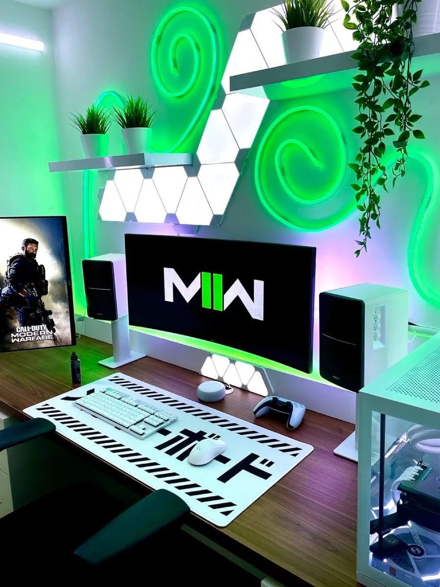 Setup gaming bernuansa hijau neon cerah dengan lampu neon fleksibel hijau yang melingkar di dinding. Terdapat monitor menampilkan logo Modern Warfare (MW), speaker putih, tanaman hias rambat, dan desk mat putih dengan aksen huruf Jepang.
