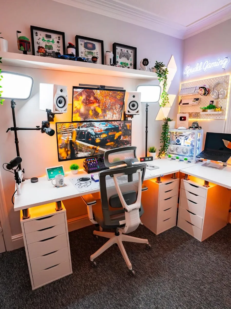 Setup PC gaming dan streaming bernuansa hangat di meja berbentuk L berwarna putih. Menampilkan dua monitor yang ditumpuk, lampu studio pencahayaan, mikrofon, kursi ergonomis abu-abu, dan rak dinding berisi koleksi gaming serta lampu neon 'Spudd Gaming'.