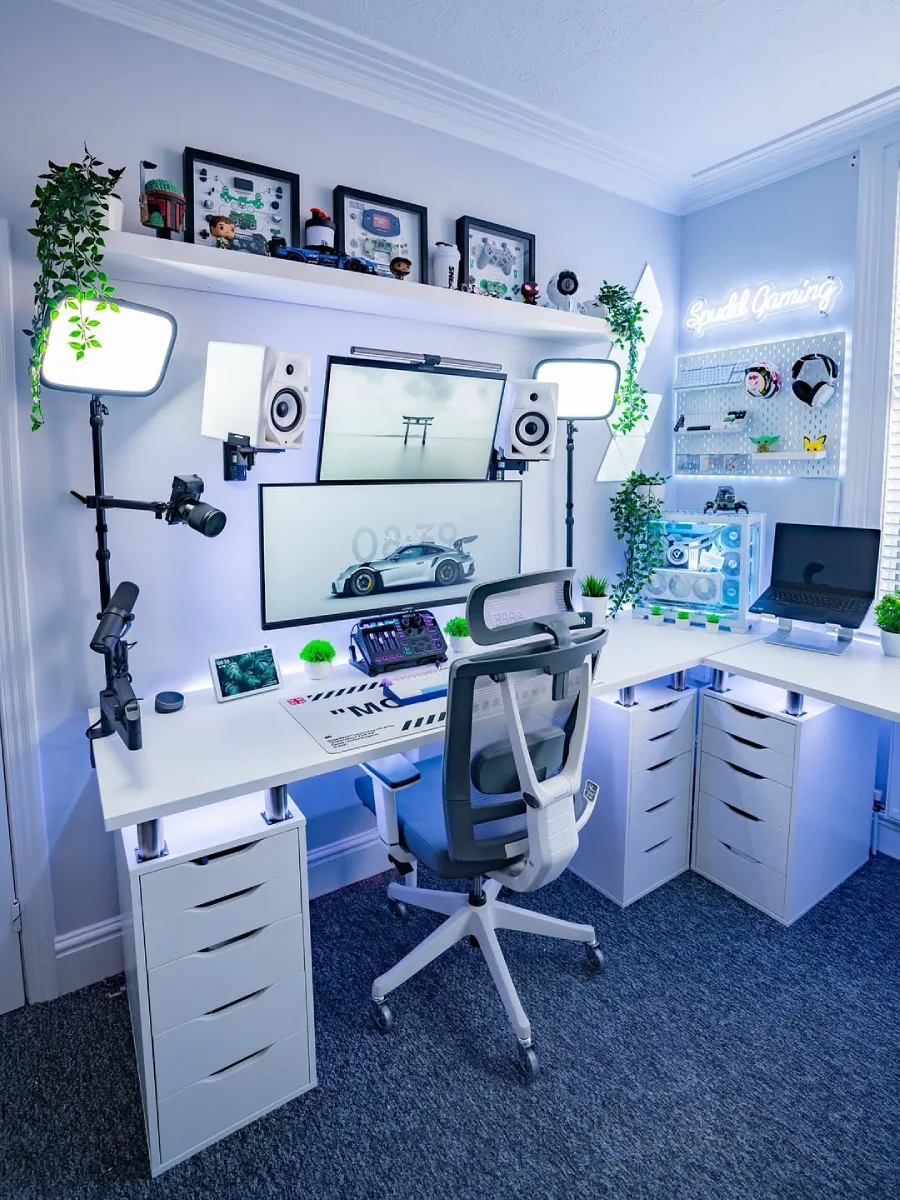 Setup PC gaming dan streaming di meja putih dengan pencahayaan biru keputihan yang sejuk. Terdapat monitor bertumpuk, sepasang lampu studio, kursi ergonomis, laptop di atas dudukan, serta rak dinding untuk aksesoris gaming.