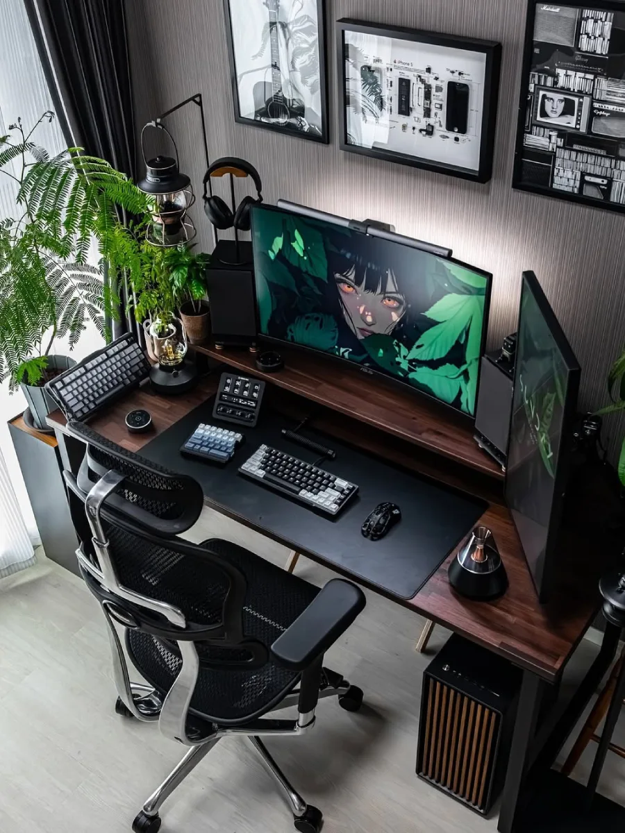 Sudut pandang atas dari setup PC gaming bernuansa gelap di atas meja kayu yang dikelilingi tanaman hias indoor. Menampilkan dua monitor menyala, keyboard mekanikal, perlengkapan audio, dan kursi ergonomis berwarna hitam.