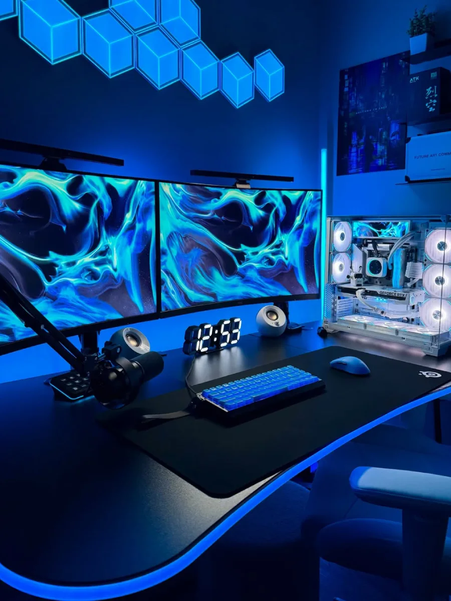 Setup PC gaming bernuansa lampu biru cerah di ruangan gelap. Menampilkan dua monitor berjejer dengan wallpaper abstrak biru, casing PC putih dengan kipas menyala, mikrofon, jam meja digital, dan lampu dinding berbentuk heksagon.