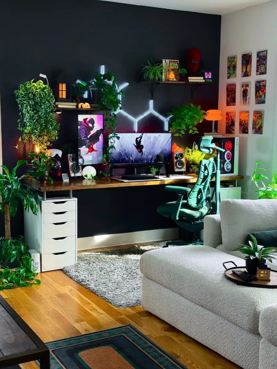 Ruang kerja bernuansa gelap dengan banyak tanaman hias merambat. Setup PC menggunakan dua monitor (satu vertikal, satu horizontal) bertema Spider-Man, lampu dinding heksagonal putih, kursi hijau, dan pajangan komik di dinding kanan. Terdapat sofa abu-abu di bagian depan.
