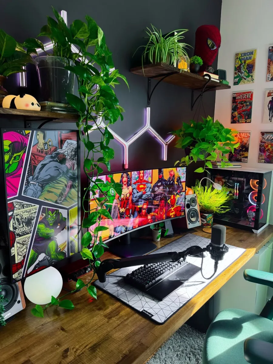 Setup PC bernuansa hangat yang dipenuhi tanaman hias daun lebar dan merambat. Menampilkan monitor lebar dengan wallpaper pahlawan super DC, poster komik besar di sebelah kiri, mikrofon di atas meja kayu, dan casing PC dengan lampu kipas RGB.