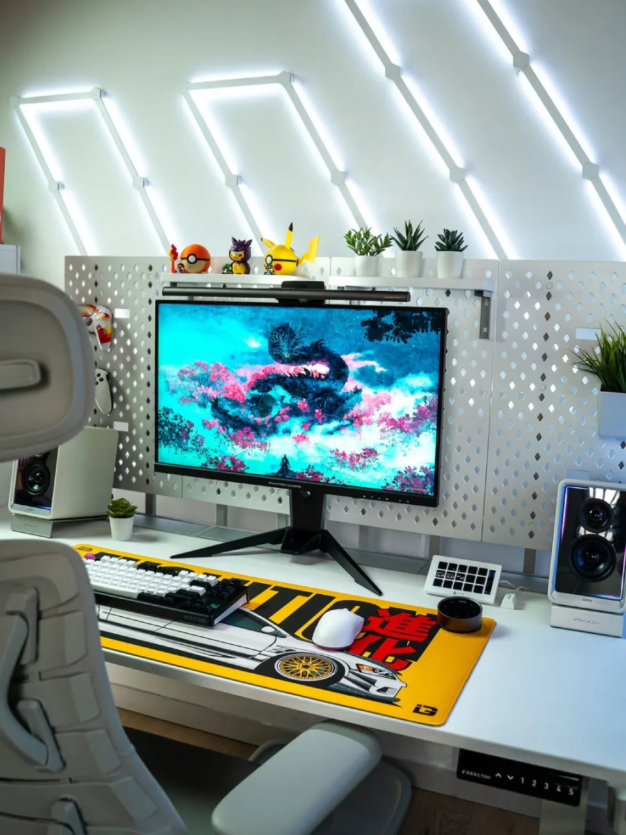 Tampilan jarak dekat dari setup PC bertema putih. Fokus pada monitor berlayar naga, desk mat kuning panjang dengan ilustrasi mobil JDM, keyboard mekanikal hitam putih, mouse putih, dan pegboard putih yang menempel di belakang meja.