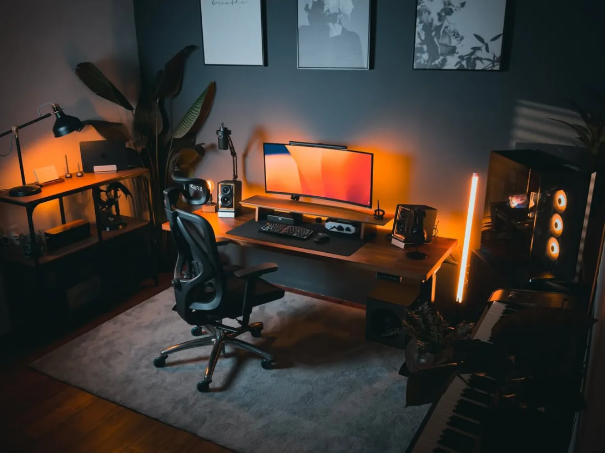 Setup home office minimalis modern dengan meja kayu, monitor widescreen, kursi ergonomis, dan lampu LED warm di ruangan bernuansa gelap.