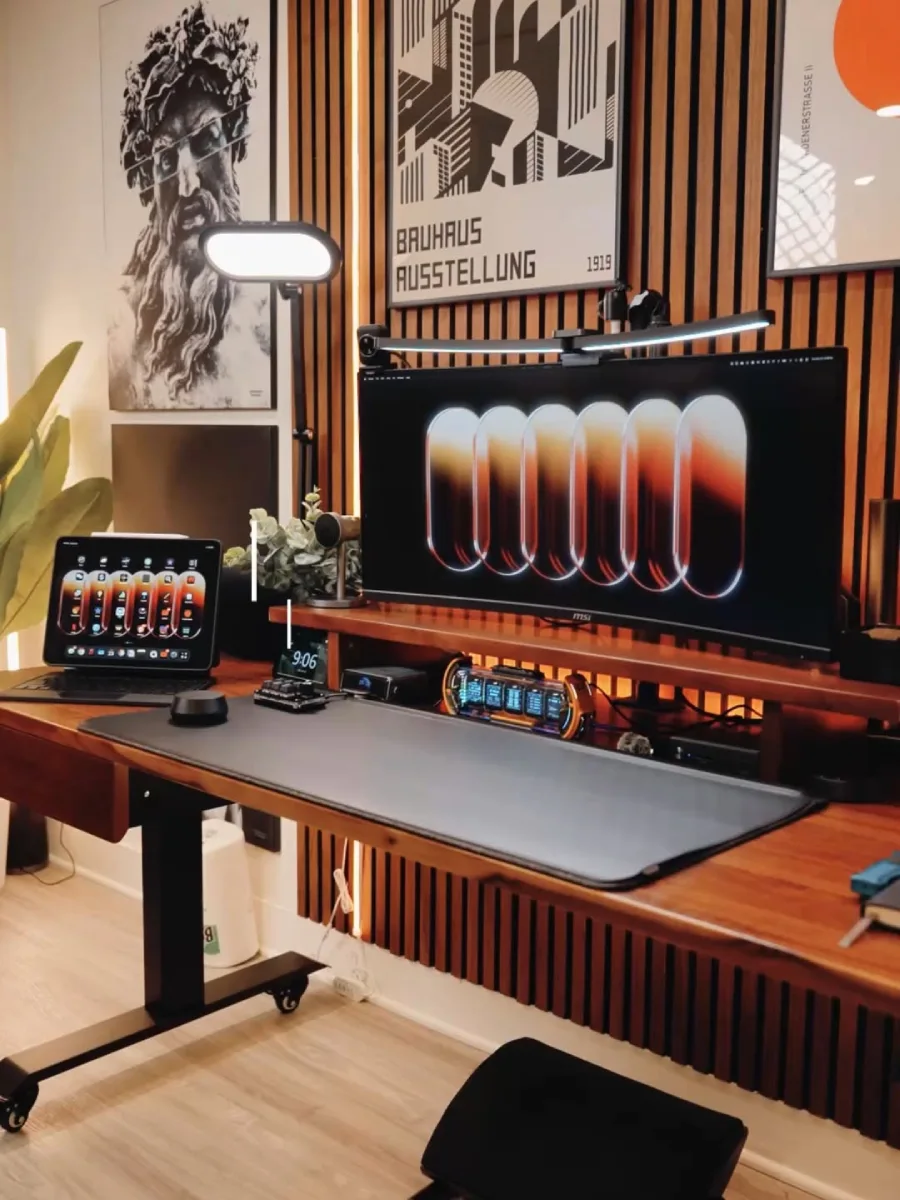 Desain home office minimalis modern dengan dinding panel kayu vertikal, meja standing desk kayu solid, monitor ultrawide, dan dekorasi poster seni klasik monokrom.