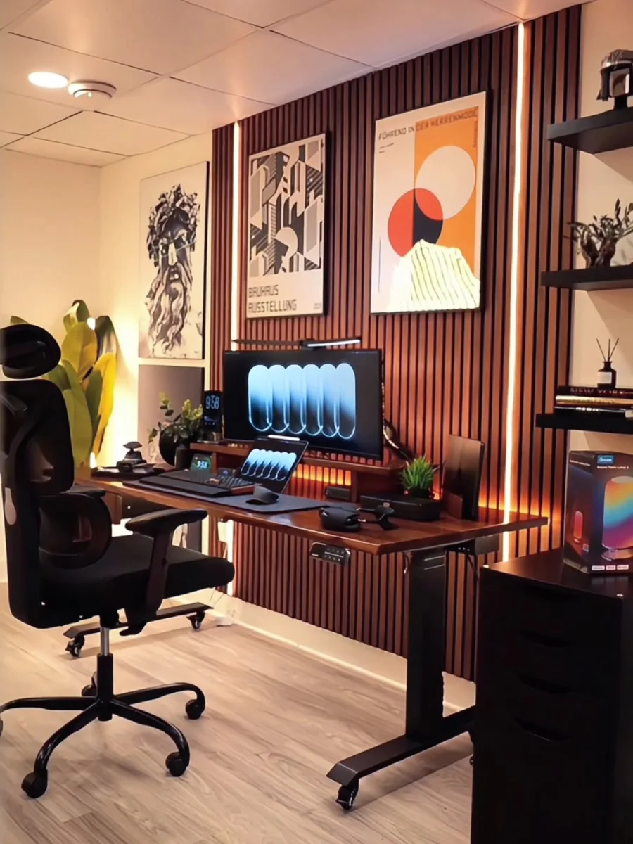 Tata ruang kerja di rumah bergaya minimalis dengan pencahayaan LED hangat di balik panel kayu, dilengkapi kursi ergonomis hitam, meja adjustable, dan rak pajangan elegan.