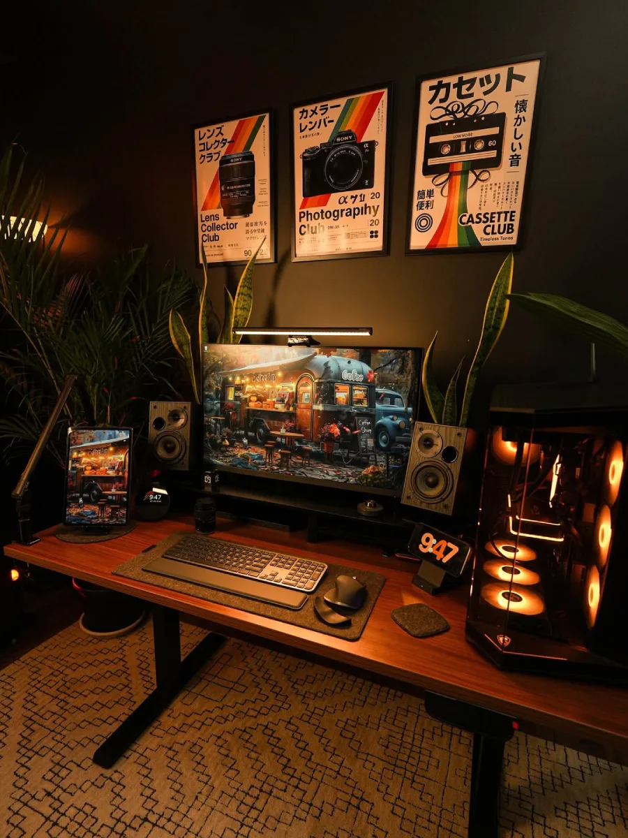 Setup meja kerja bernuansa gelap yang maskulin, menampilkan cahaya ambient oranye, poster retro Jepang di dinding hitam, PC desktop, dan tanaman hias indoor.