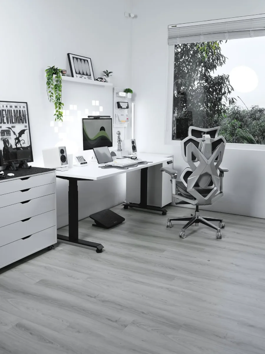Ruang kerja minimalis bernuansa putih dan abu-abu yang dilengkapi meja kerja berdiri, kursi ergonomis berdesain futuristik, dan jendela besar menghadap pepohonan hijau.