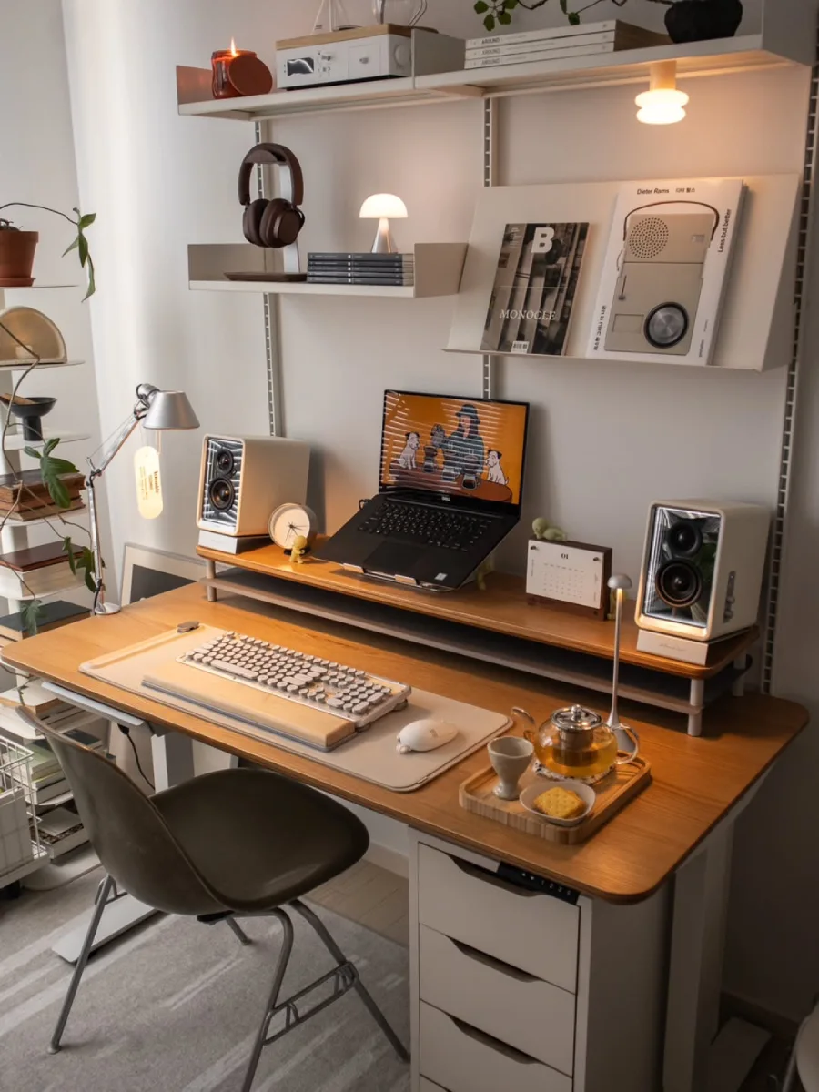 Tampilan sudut samping meja kerja minimalis modern bernuansa kayu dan putih. Menyoroti detail di atas meja seperti keyboard mekanikal dengan sandaran tangan kayu, set teh hangat dan camilan di atas nampan kecil, serta laptop yang diletakkan pada dudukan ergonomis di atas rak tambahan. Terlihat juga detail laci penyimpanan berwarna putih di bawah meja bagian kanan.