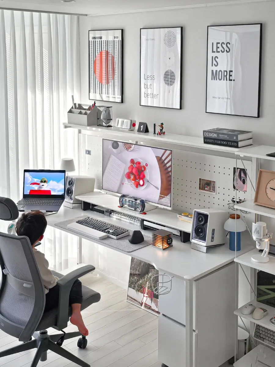 Ruang kerja serba putih yang terang benderang dengan cahaya alami, meja lebar, kursi ergonomis abu-abu, dan pegboard putih untuk pengorganisasian barang.